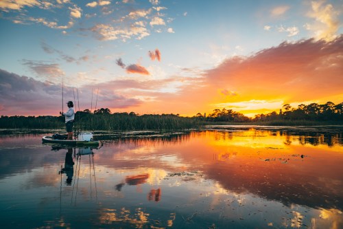 A person fishing on a calm lake at sunset, with vibrant colors reflecting on the water and lush greenery in the background.