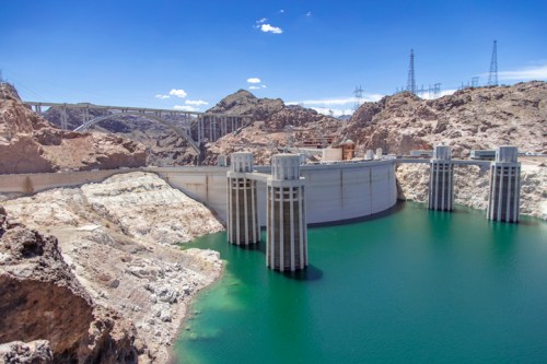 A large dam with towering structures, surrounded by rocky terrain and a turquoise reservoir under a clear blue sky.