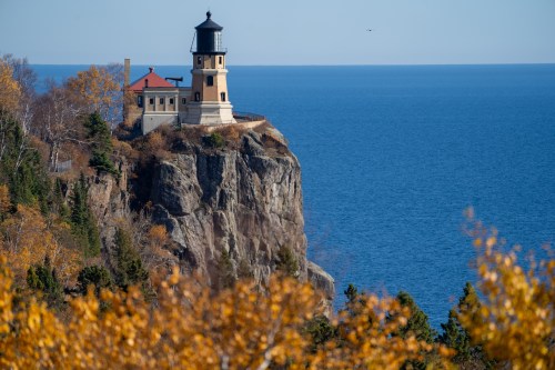 A lighthouse perched on a rocky cliff overlooking a calm blue lake, surrounded by autumn foliage.