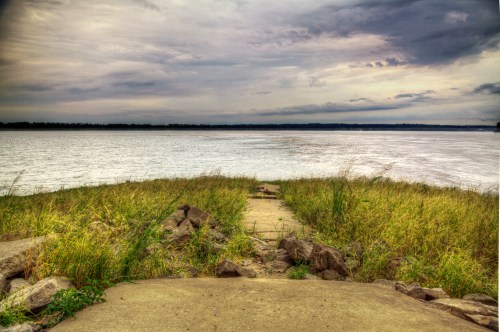 A grassy path leads to a calm river under a cloudy sky, with rocks lining the walkway.