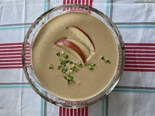 A bowl of creamy soup topped with apple slices and chopped green onions, set on a checkered tablecloth.