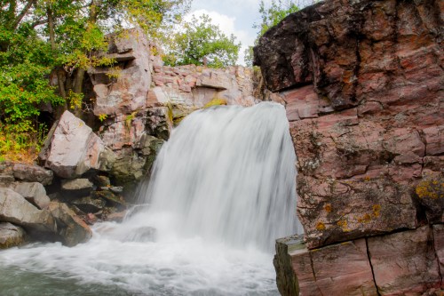 A cascading waterfall flows over rocky cliffs, surrounded by lush greenery and a bright blue sky.
