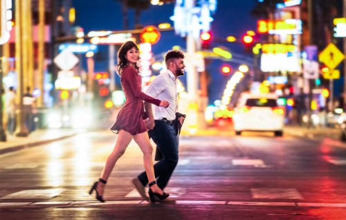 A couple joyfully crossing a busy street at night, surrounded by colorful city lights and traffic.