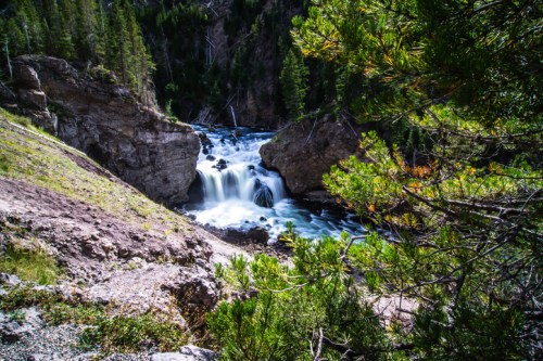 A serene waterfall cascades through rocky terrain, surrounded by lush green trees and a vibrant natural landscape.