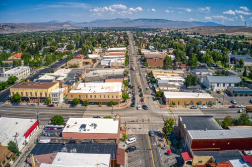 Aerial view of a small town with a main street lined by shops, trees, and mountains in the background under a clear blue sky.