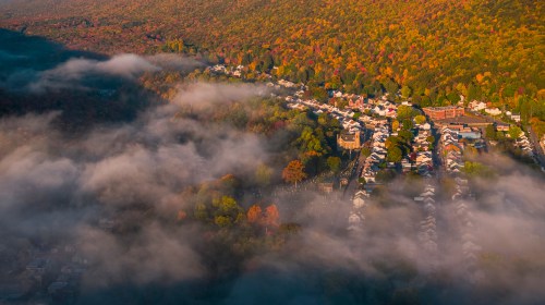 Aerial view of a colorful autumn landscape with fog over a town surrounded by vibrant fall foliage.