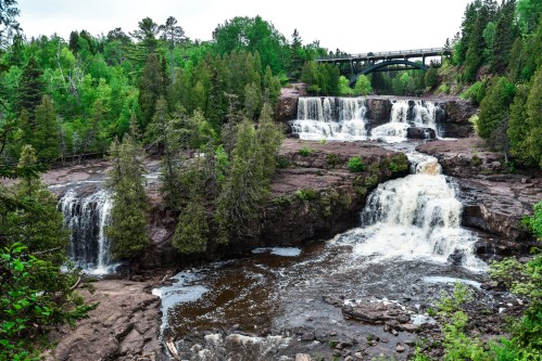 A scenic view of cascading waterfalls surrounded by lush greenery and a bridge in the background.
