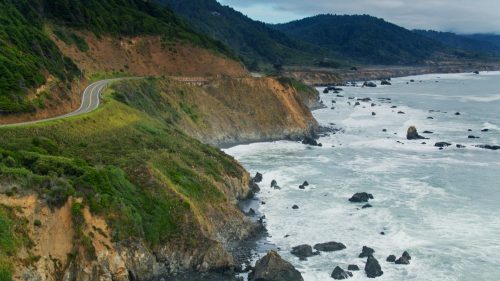 A scenic coastal view with a winding road along cliffs, waves crashing against rocky shores, and lush green hills in the background.