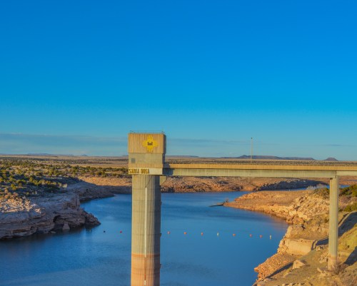 A concrete bridge spans a calm blue lake under a clear blue sky, with rocky shores visible in the background.