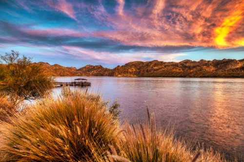 A serene river scene at sunset, with colorful clouds, mountains in the background, and lush grasses in the foreground.