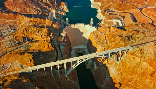 Aerial view of a large dam surrounded by rocky terrain and a river, with a bridge crossing above it.