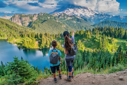 Two children stand on a trail overlooking a lush green landscape and a mountain, pointing at the view.