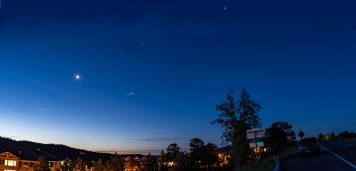 A panoramic night sky with stars and a crescent moon above a quiet roadside and distant buildings.