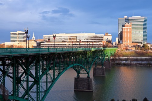 A green bridge spans a river, with modern buildings and a cloudy sky in the background.