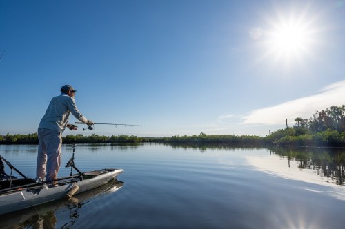 A person fishing from a kayak on a calm river, with lush greenery and a bright sun in the clear blue sky.