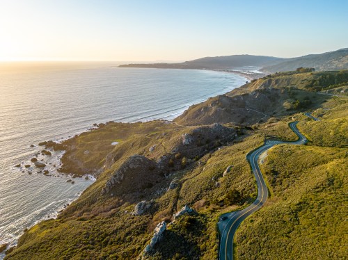 Aerial view of a winding coastal road along a scenic shoreline at sunset, with hills and ocean waves in the background.