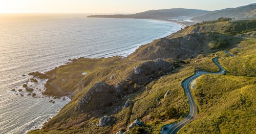 Aerial view of a coastal landscape with winding roads, green hills, and the ocean at sunset.