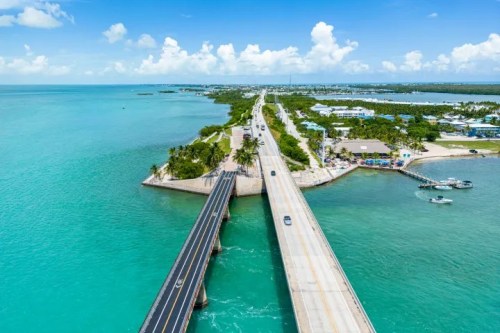 Aerial view of a coastal highway splitting into two bridges over turquoise waters, surrounded by greenery and boats.