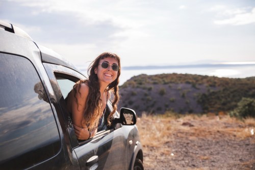 A smiling woman with sunglasses leans out of a car window, enjoying a scenic view of hills and water.