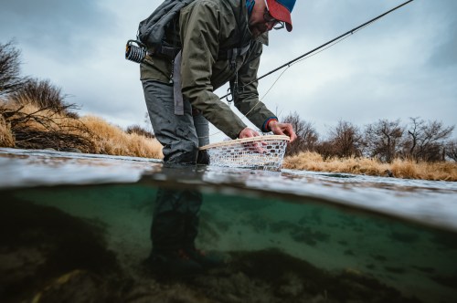 A person in fishing gear kneels in shallow water, using a net to catch fish, with a cloudy sky in the background.