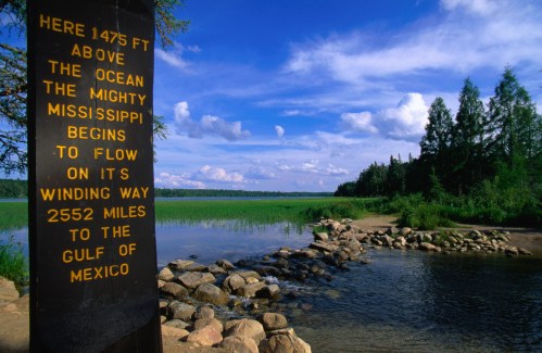 A sign marking the start of the Mississippi River, with a scenic view of water and trees in the background.