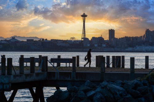 Silhouette of a person walking on a pier at sunset, with the Seattle skyline and Space Needle in the background.