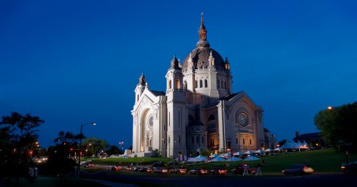 A grand cathedral illuminated at dusk, surrounded by trees and parked cars, with a clear blue sky above.