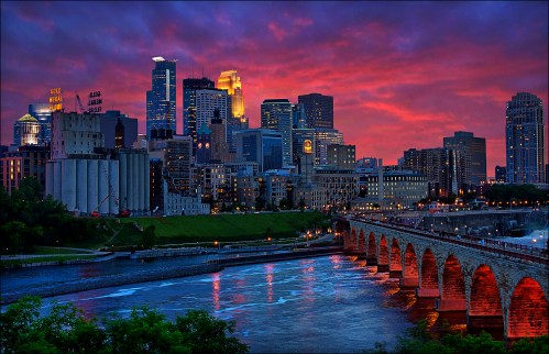A vibrant sunset over a city skyline, reflecting on a river with a historic stone bridge in the foreground.