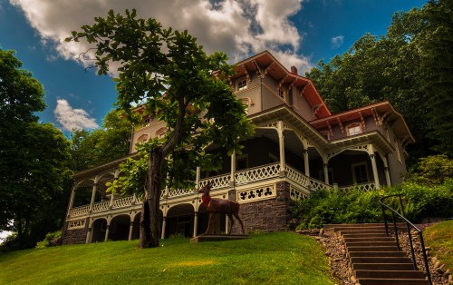 A large, historic house with a wraparound porch, surrounded by trees and greenery under a partly cloudy sky.