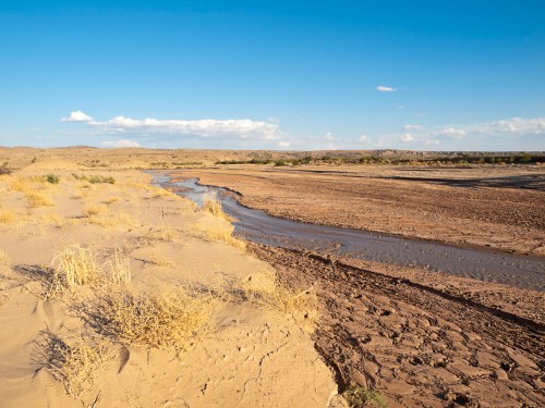A dry desert landscape with sparse vegetation and a winding stream under a blue sky with scattered clouds.