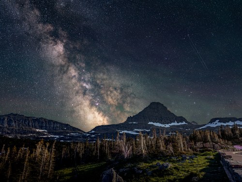 A starry night sky with the Milky Way over a mountain landscape, featuring snow and trees in the foreground.