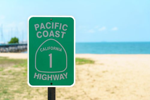 Green sign for Pacific Coast Highway 1, with a beach and ocean in the background under a clear blue sky.