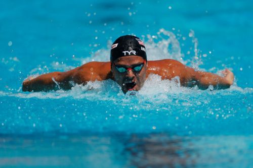 A swimmer performs the butterfly stroke in a bright blue pool, creating splashes of water around him.