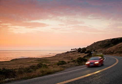 A red car speeds along a coastal road at sunset, with ocean waves and hills in the background.