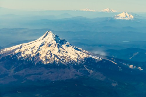 Aerial view of a snow-capped mountain peak surrounded by rolling hills and distant mountains under a clear sky.
