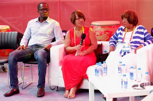 Three people seated on a stage, engaged in discussion, with water bottles and traditional drums in the background.