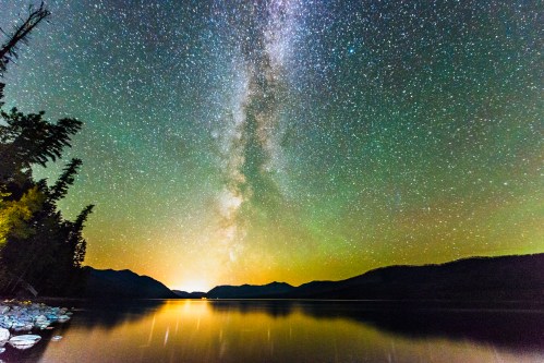A stunning night sky filled with stars and the Milky Way, reflecting on a calm lake surrounded by mountains and trees.