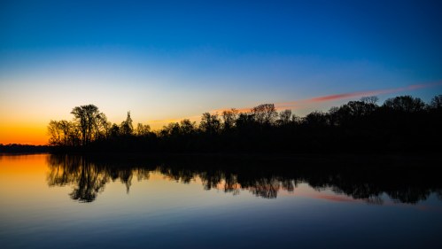 A serene sunset over a calm lake, with silhouetted trees reflecting on the water's surface.