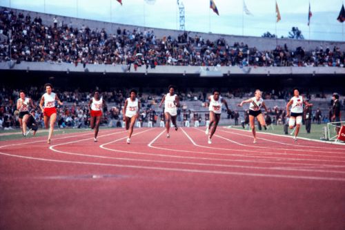 A group of female athletes competing in a race on a track, with a cheering crowd in the background.