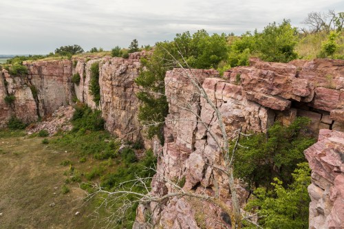 A rocky cliff with pink and gray stone, surrounded by greenery and a cloudy sky in the background.