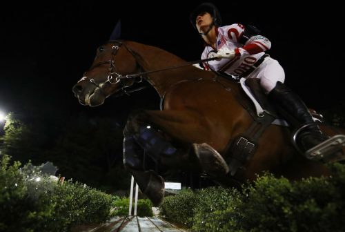 A rider in equestrian gear jumps over an obstacle on a horse at night, illuminated by bright lights.