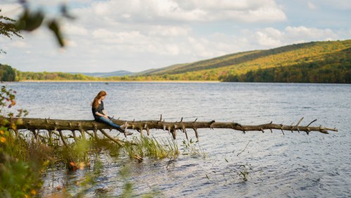 A woman sits on a fallen tree by a serene lake, surrounded by colorful autumn foliage and rolling hills.