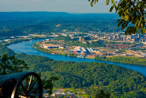Aerial view of a river winding through green hills, with industrial buildings and a city in the distance under a clear sky.