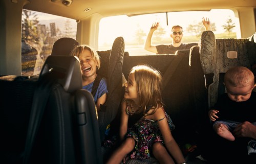 Two children laugh in a car while a man waves from outside. A baby sits quietly in a car seat. Sunlight streams in.