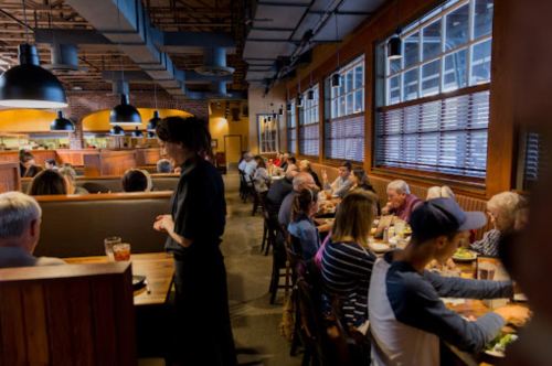 A busy restaurant interior with diners at tables and a server attending to guests. Warm lighting and wooden decor.