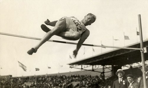 A high jumper clears the bar during a competition, with spectators and flags in the background.