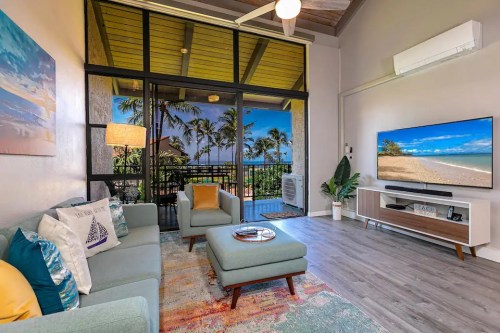 Bright living room with modern furniture, large windows, and a view of palm trees and a beach outside.