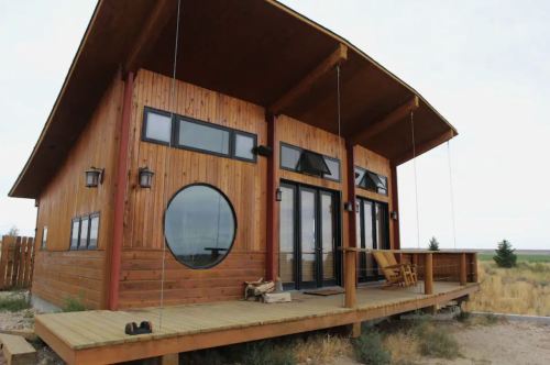 A modern wooden house with large windows and a circular window, featuring a deck and a scenic outdoor view.