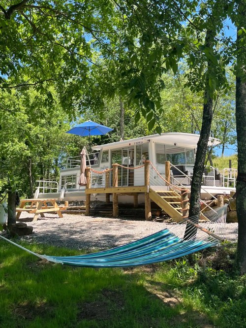 A cozy cabin surrounded by trees, featuring a deck, hammock, and blue umbrella for shade on a sunny day.