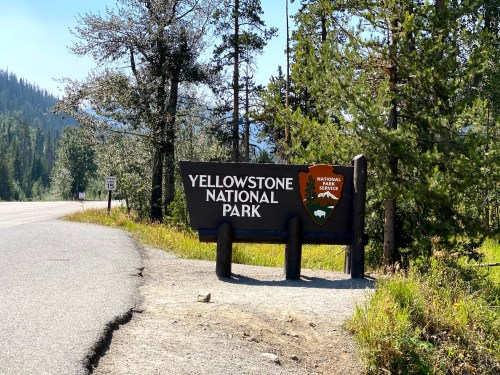 Sign for Yellowstone National Park along a road, surrounded by trees and mountains in the background.